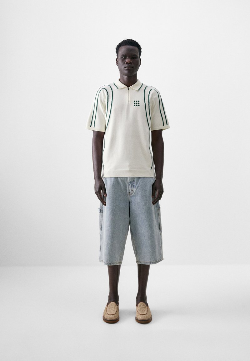 Beige knitted polo shirt with green vertical stripes, paired with blue denim cargo shorts. Woven slip-on shoes complete the outfit.