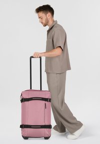 Rosa duffelväska med svarta detaljer, rektangulär form, dragkedjestängning och justerbara remmar. Slät tygtextur.