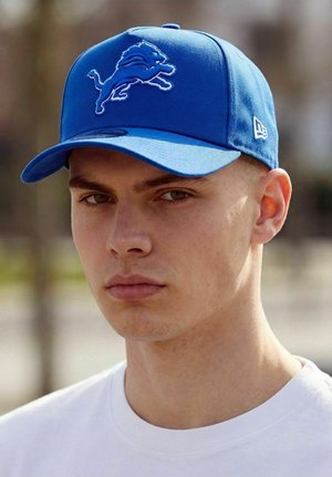 Young man wearing a blue baseball cap with a lion logo and a white shirt, outdoors in soft daylight.