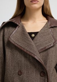 Cappotto in tweed marrone con colletto in pelle marrone, design doppiopetto e un accento di bottone dorato al collo. Tessuto testurizzato visibile.