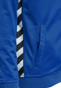 Blå sportjacka med texturerat tyg, sidoficka och randig detaljer i svart och vitt på insidan.