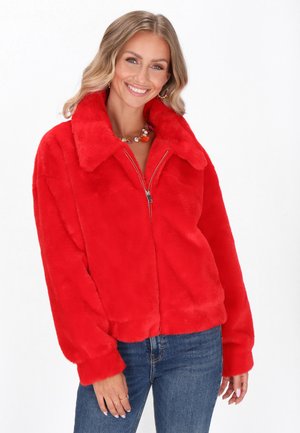 O femeie zâmbitoare cu păr ondulat blond poartă o jachetă roșie din blană synthetică cu fermoar și blugi, fiind accessoruită cu o bijuterie colorată la gât și cercei.