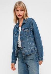 Jovem mulher com cabelo loiro, vestindo uma jaqueta de denim azul e jeans sobre uma blusa branca, de pé contra um fundo claro e liso.
