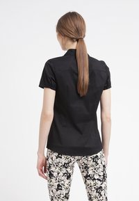 Camicia nera a maniche corte con un design aderente, dotata di un yoke posteriore e dettagli con bottoni. Abbinata a pantaloni con stampa floreale.