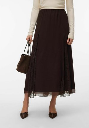 Femme portant une longue jupe marron foncé avec des panneaux en dentelle, un haut crème à manches longues, des ballerines marron pointues cloutées, et tenant un sac à main marron.