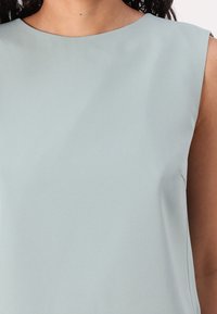 Donna che indossa una canotta azzurra chiara con scollo rotondo su uno sfondo uniforme, che mostra il busto superiore e parte del braccio.