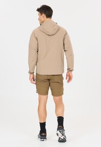 Homme portant une veste à capuche beige, un short marron, des chaussettes noires et des chaussures de course, debout et tourné vers l'arrière sur un fond blanc.