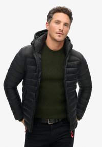 Giacca a vento nera con cappuccio, fronte con zip e cuciture texturizzate. Indossata sopra un maglione a maglia verde scuro. Notevole accentuazione rossa della zip.