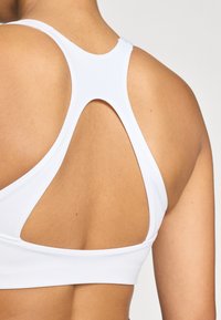 Fehér sportmelltartó versenyháttal és kivágott részlettel. Simább, rugalmas anyag, amely kényelmesen illeszkedik a testhez.