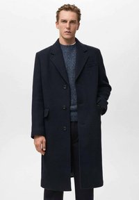 Cappotto sopra il ginocchio in lana blu navy con design a un solo petto, ampi rever, quattro bottoni frontali e tasche laterali. Indossato sopra un maglione a trama.