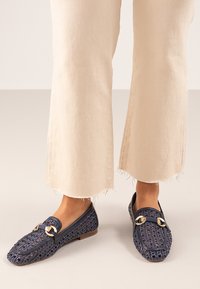 Mocasines de tela azul marino con detalles en dorado y un acabado texturizado, combinados con pantalones cortos de color beige claro.