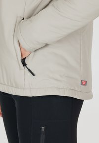 Vaaleanbeige puff jacket, jossa on vetoketjullinen tasku vasemmalla puolella, elastiset helmat ja pieni punainen brändilogo lähellä alareunaa.