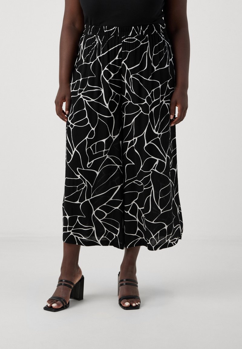 Personne portant un pantalon noir à jambes larges avec des motifs de lignes abstraites blanches et des sandales noires à talons avec double bride.