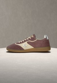 Vue latérale d'une basket basse bordeaux et beige avec des lacets blancs, une semelle en gomme et une languette métallique argentée au talon.