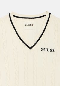 Πουλόβερ με V λαιμόκοψη σε κρεμ απόχρωση, με σχέδιο πλεξούδας, μαύρη ριμπ λαιμόκοψη και μαύρη κεντημένη στάμπα "GUESS".