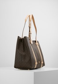 Borsa tote sintetica marrone con accenti crema e strisce verticali, caratterizzata da un motivo a monogramma e manici in pelle piatti. La base ha piedi in metallo.