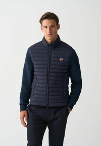 Navy puffer vest features horizontal stitching, high collar, and two side pockets. Long sleeves with a solid navy fabric. Logo on chest.