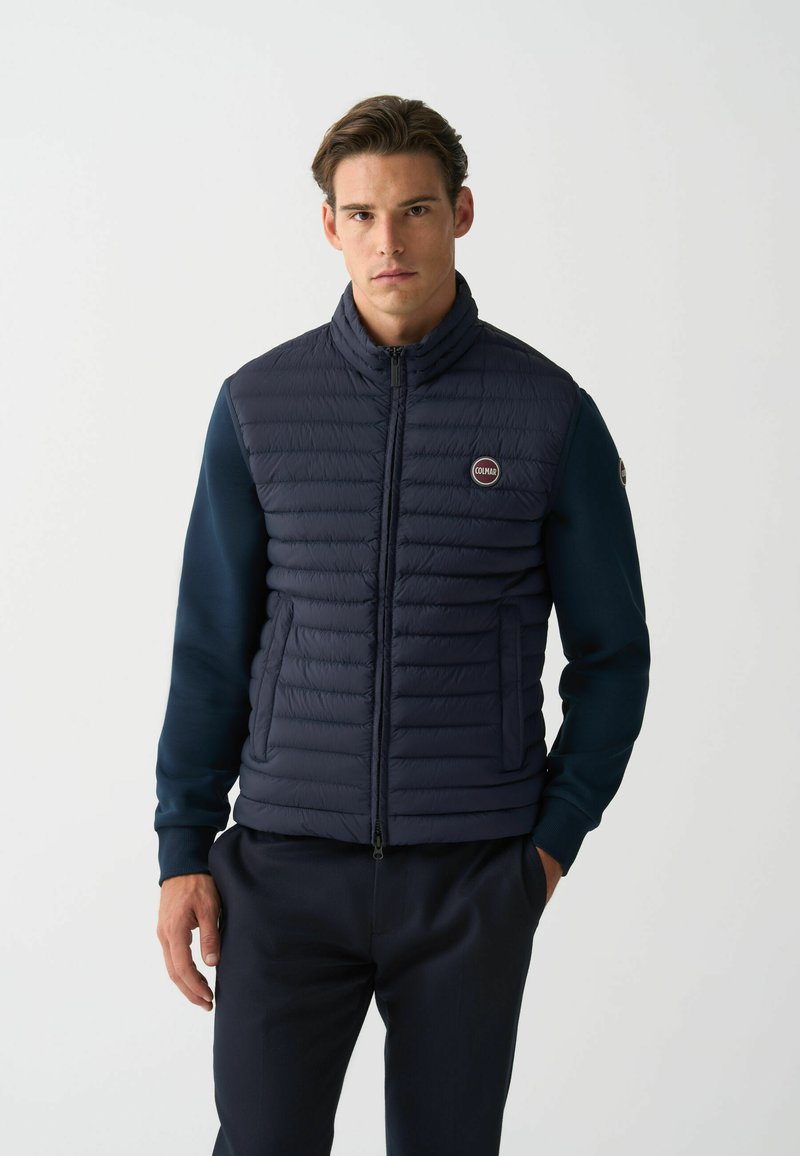 Navy puffer vest features horizontal stitching, high collar, and two side pockets. Long sleeves with a solid navy fabric. Logo on chest.