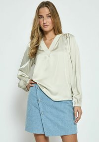 Blusa satin beige chiaro con scollatura a V e maniche a sbuffo, abbinata a una gonna blu con texture e chiusura laterale a bottone.