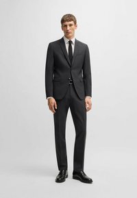 Dunkelgrau Anzug mit maßgeschneiderter Passform, einreihiger Jacke, Reverskragen und passender Hose; kombiniert mit einem weißen Hemd und einer schwarzen Krawatte.