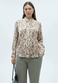 Beige slangenprint blouse met een kraaghalslijn en lange mouwen, gemaakt van gladde stof. Gecombineerd met lichtgrijze broeken en een kleine zwarte tas.