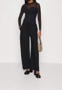 Svart långärmad topp i genomskinligt material med prickiga detaljer, ihop med högmidjade, vida svarta byxor och kraftiga svarta skor.