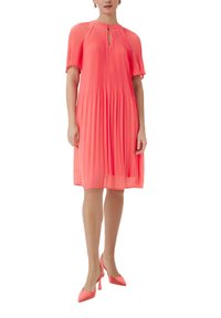 Robe plissée, longueur genou, d'un corail éclatant, avec un décolleté en trou de serrure et des manches courtes. Associée à des talons hauts corail assortis.