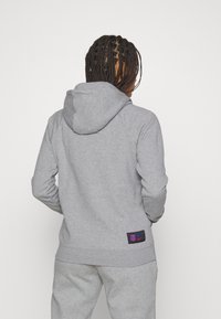 Graue Kapuzenpulli aus weichem Stoff mit Kapuze, ausgestattet mit einem Markenaufnäher in Blau und Pink an der unteren linken Ecke. Einfaches Design, ohne Muster.