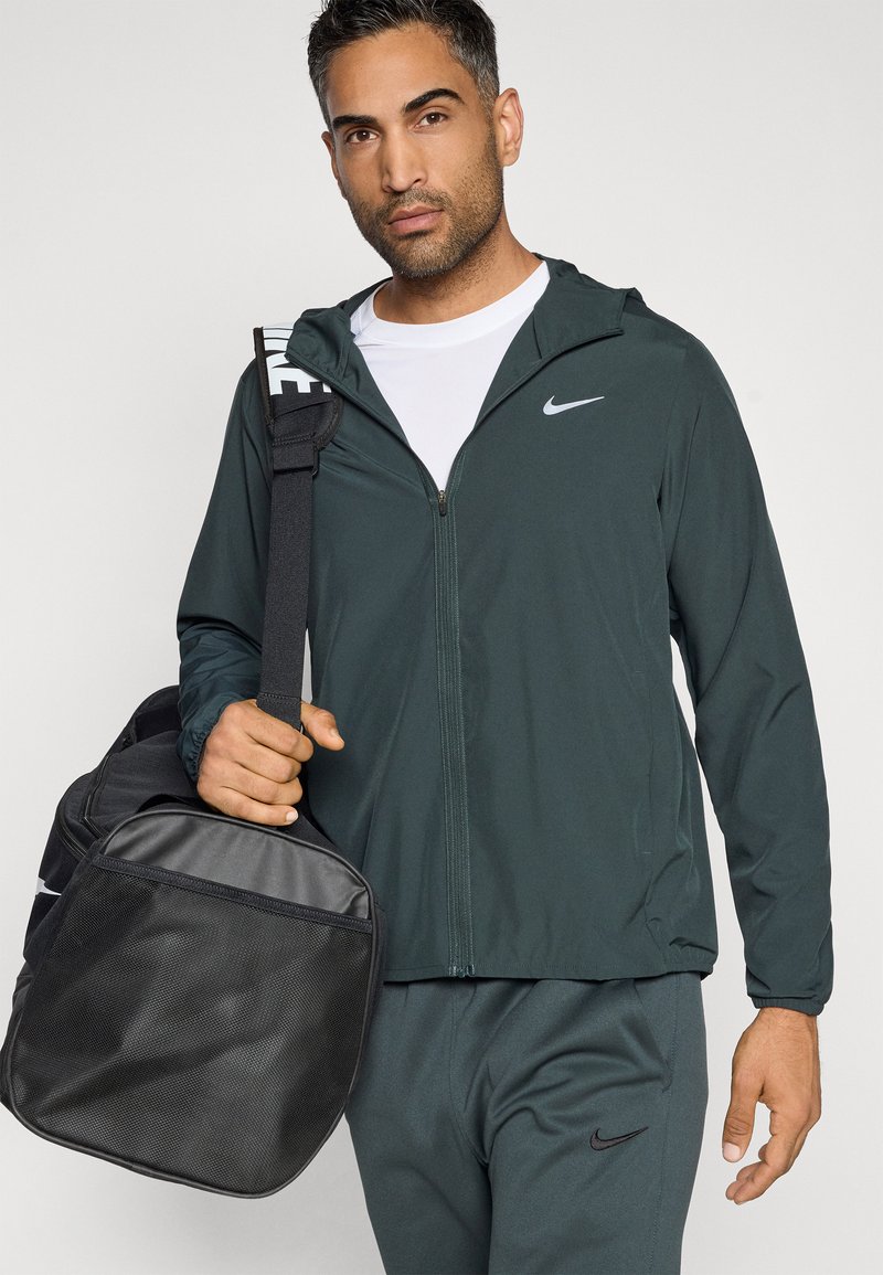 Chaqueta ligera de color verde oscuro con capucha, cierre frontal y logo de Nike en blanco, combinada con pantalones a juego; lleva un bolso de duffel negro texturizado.