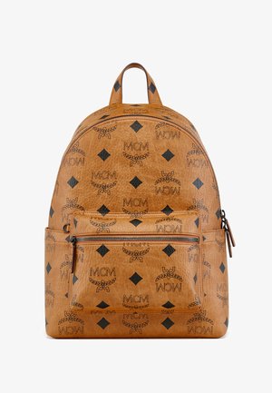 Mochila de couro sintético castanho com superfície texturizada, padrão proeminente de logótipos MCM em preto, compartimentos duplos com fechos e correias ajustáveis.