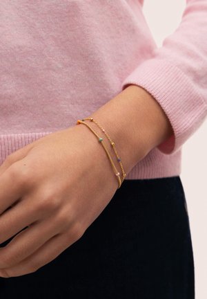 Muñeca con dos delicadas pulseras de oro, una con pequeñas cuentas de colores, lucidas sobre un suéter de manga larga rosa y pantalones oscuros.