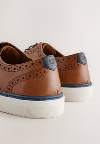 Braune Lederschuhe mit detaillierten Perforationen und marineblauen Akzenten. Weiße Gummisohle mit strukturierten Streifen und einem Logo an der Ferse.