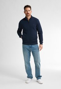 Námořnická mikina na zip s žebrovanými manžetami a lemem, texturovaná látka, nošená přes šedé tričko, v kombinaci se světle modrými džíny a bílými teniskami.