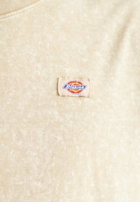 Camiseta de algodón color crema con una sutil textura veteada, que presenta un parche de logo tejido de Dickies en el pecho.