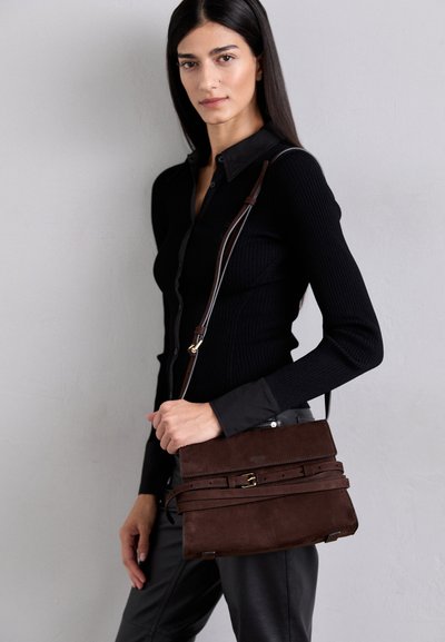 Bolso de mano de ante marrón con una superficie texturizada, que cuenta con una tapa frontal asegurada por una correa decorativa y una larga correa de hombro ajustable.