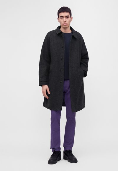 Man wearing dark gray buttoned coat, black shirt, purple pants, and black shoes standing with one hand in pocket against plain background.