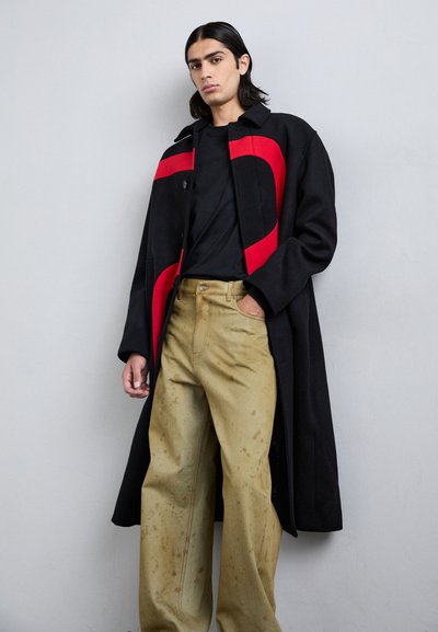 Cappotto di lana nero con accenti curvi rossi, silhouette lunga, indossato su una t-shirt nera e pantaloni chiari color khaki con una trama sbiadita.