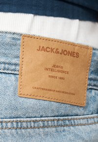 Δερμάτινο πατς σε ανοιχτόχρωμο μπλε τζιν με το κείμενο «JACK & JONES», «JEANS INTELLIGENCE ΑΠΟ ΤΟ 1990» και «ΤΕΧΝΗ ΚΑΙ ΑΦΙΕΡΩΣΗ.»