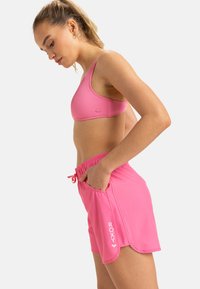 Roze sportbeha en korte broek gemaakt van een gladde, rekbare stof. De beha heeft een geribbelde textuur; de korte broek heeft zijzakken en een verstelbare taille.