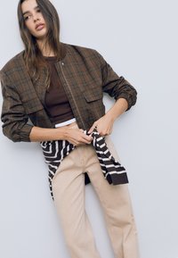 Smeđi karirani bomber jakna s elastičnim manžetama, nosi se preko tamnog gornjeg dijela. Bež hlače, prugasti pulover vezan u struku, neutralna pozadina.