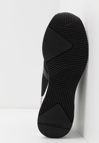 Suela de zapato de goma negra con patrones de agarre texturizados. Presenta un diseño angular y la marca "MICHAEL KORS" grabada en la parte inferior.