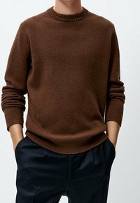 Brązowy sweter dziany z prążkowanym okrągłym dekoltem i długimi rękawami, zestawiony z ciemnymi spodniami, charakteryzującymi się luźnym krojem i gładką teksturą.