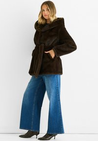 Brown faux fur coat with a wide collar, belted waist, and side pockets, worn over light blue wide-leg jeans and snake print heels.