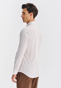 Biała koszula w małe niebieskie, czerwone i czarne kropki, z długimi rękawami i kołnierzykiem zapinanym, w zestawie z brązowymi spodniami. Gładka faktura materiału.