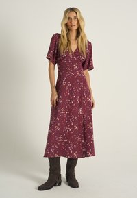 Rochie maro roșcat cu mâneci scurte evazate și decolteu în V. Materialul are un model mic, floral și colorat. Asortată cu cizme maro până la genunchi.