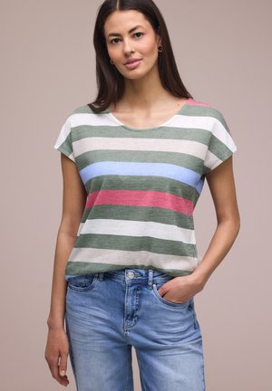 Junge Frau mit langen, dunklen Haaren, die ein kurzärmliges gestreiftes Hemd in Grün, Weiß, Blau und Rot sowie eine hellblaue Jeans trägt, mit einer Hand in der Tasche.