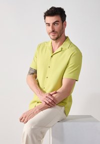 Camisa de manga corta, texturizada, de color verde claro con cuello y botones, combinada con pantalones beige. El modelo está sentado sobre un pedestal de color claro.