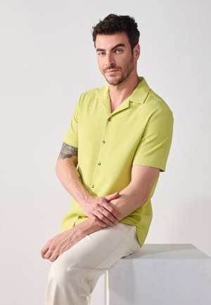 Camisa de manga corta, texturizada, de color verde claro con cuello y botones, combinada con pantalones beige. El modelo está sentado sobre un pedestal de color claro.