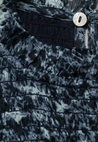 Tessuto scuro a fantasia con una texture a pieghe, dotato di un pulsante bianco rotondo al collo. Il design include diverse tonalità di blu e nero.