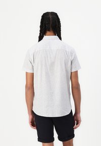 Camicia a maniche corte in tessuto leggero, bianca con un motivo a piccoli punti neri, dotata di colletto standard e dettagli sulla spalla posteriore.